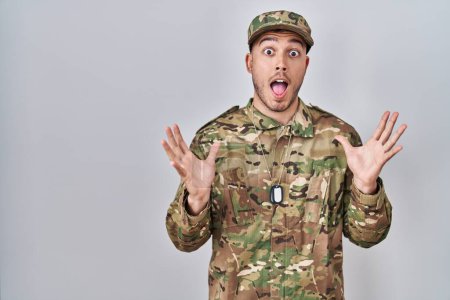 Foto de Joven hombre hispano vistiendo uniforme camuflaje del ejército celebrando loco y sorprendido por el éxito con los brazos levantados y los ojos abiertos gritando emocionado. concepto ganador - Imagen libre de derechos