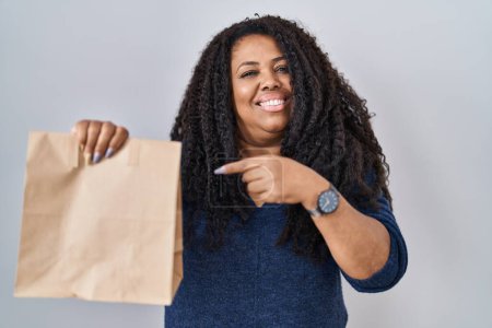 Téléchargez les photos : Plus taille hispanique femme tenant emporter sac en papier souriant heureux pointage avec la main et le doigt - en image libre de droit