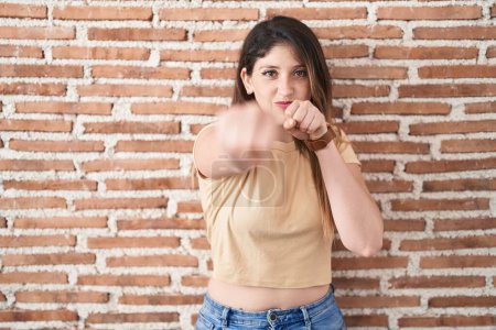 Téléchargez les photos : Jeune femme brune debout sur un mur de briques poing poing pour se battre, attaque agressive et en colère, menace et violence - en image libre de droit