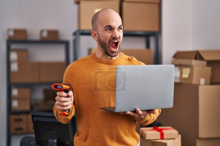 Photo for Young bald man with beard working at small business ecommerce with laptop angry and mad screaming frustrated and furious, shouting with anger. rage and aggressive concept. - Royalty Free Image