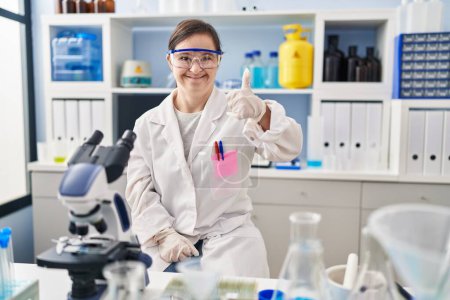 Téléchargez les photos : Hispanic girl with down syndrome working at scientist laboratory doing happy thumbs up gesture with hand. approving expression looking at the camera showing success. - en image libre de droit