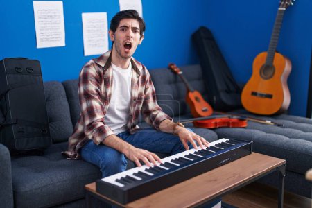 Foto de Joven hispano tocando el piano en el estudio de música enojado y loco gritando frustrado y furioso, gritando con ira. rabia y concepto agresivo. - Imagen libre de derechos