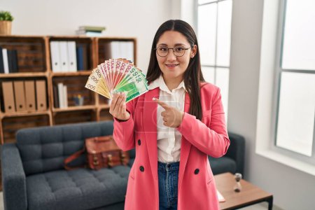 Téléchargez les photos : Jeune femme latine travaillant au bureau de consultation tenant de l'argent souriant heureux pointant avec la main et le doigt - en image libre de droit