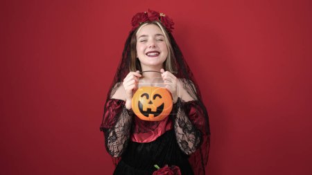Téléchargez les photos : Jeune femme blonde portant katrina costume tenant panier de citrouille halloween sur fond rouge isolé - en image libre de droit