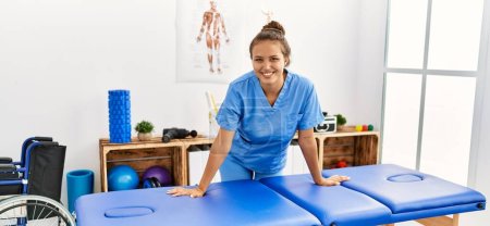 Foto de Joven hermosa mujer hispana fisioterapeuta sonriendo confiada de pie en la clínica de rehabilitación - Imagen libre de derechos