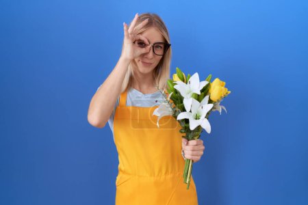 Foto de Mujer joven caucásica con delantal de florista sosteniendo flores haciendo buen gesto con la mano sonriente, ojos mirando a través de los dedos con la cara feliz. - Imagen libre de derechos