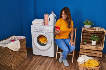 Photo for Young latin woman using smartphone waiting for washing machine at laundry room - Royalty Free Image