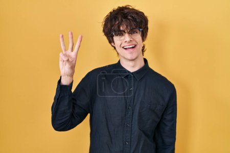 Foto de Joven con gafas sobre fondo amarillo mostrando y apuntando hacia arriba con los dedos número tres mientras sonríe confiado y feliz. - Imagen libre de derechos