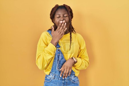 Téléchargez les photos : Femme africaine debout sur fond jaune ennuyé bâillement fatigué couvrant la bouche avec la main. agitation et somnolence. - en image libre de droit