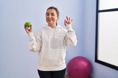 Foto de Mujer hispana de mediana edad con ropa deportiva sosteniendo manzana sana haciendo signo de ok con los dedos, sonriendo gesto amistoso excelente símbolo - Imagen libre de derechos