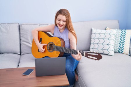 Photo for Young redhead woman having online classical guitar class sitting on sofa at home - Royalty Free Image