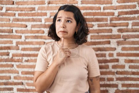 Téléchargez les photos : Jeune femme hispanique debout sur le mur de briques pensée concentrée sur le doute avec le doigt sur le menton et la recherche jusqu'à se demander - en image libre de droit