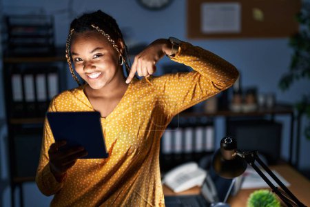 Photo for African american woman with braids working at the office at night with tablet looking confident with smile on face, pointing oneself with fingers proud and happy. - Royalty Free Image