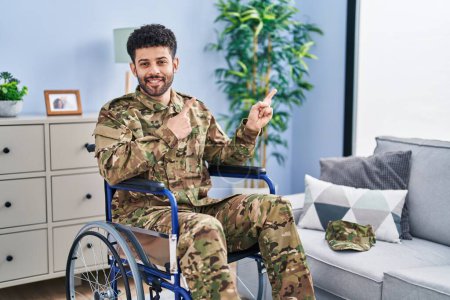 Foto de Hombre árabe vistiendo uniforme camuflaje del ejército sentado en silla de ruedas sonriendo y mirando a la cámara apuntando con dos manos y dedos hacia un lado. - Imagen libre de derechos