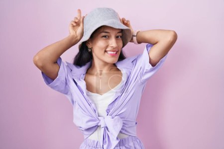 Foto de Mujer hispana joven de pie sobre fondo rosa con sombrero posando divertido y loco con los dedos en la cabeza como orejas de conejo, sonriente alegre - Imagen libre de derechos