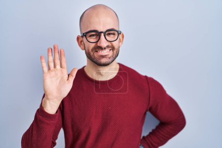 Foto de Joven hombre calvo con barba de pie sobre fondo blanco con gafas renunciando a decir hola feliz y sonriente, gesto de bienvenida amistoso - Imagen libre de derechos