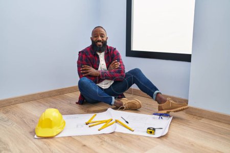 Photo for African american man sitting on the floor at new home looking at blueprints happy face smiling with crossed arms looking at the camera. positive person. - Royalty Free Image