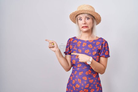 Foto de Mujer caucásica joven con vestido de flores y sombrero de verano señalando a un lado preocupado y nervioso con ambas manos, expresión preocupada y sorprendida - Imagen libre de derechos