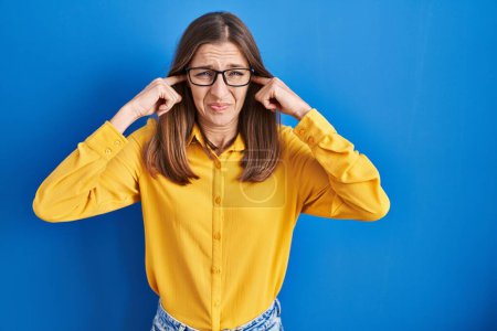 Téléchargez les photos : Young woman wearing glasses standing over blue background covering ears with fingers with annoyed expression for the noise of loud music. deaf concept. - en image libre de droit
