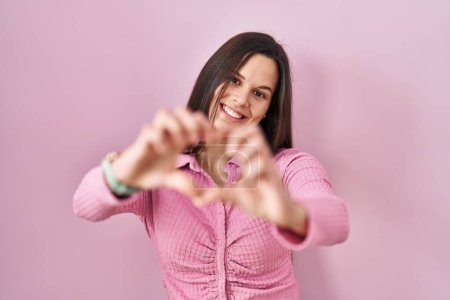Foto de Young hispanic woman standing over pink background smiling in love doing heart symbol shape with hands. romantic concept. - Imagen libre de derechos