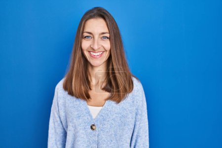 Foto de Mujer joven de pie sobre fondo azul con una sonrisa alegre y fresca en la cara. persona afortunada. - Imagen libre de derechos