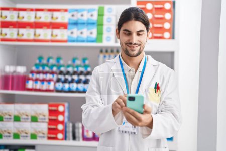 Téléchargez les photos : Jeune homme hispanique pharmacien utilisant un smartphone travaillant à la pharmacie - en image libre de droit