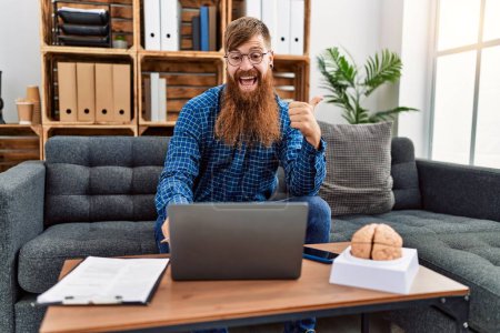 Photo for Redhead man with long beard using laptop working at psychology clinic pointing thumb up to the side smiling happy with open mouth - Royalty Free Image