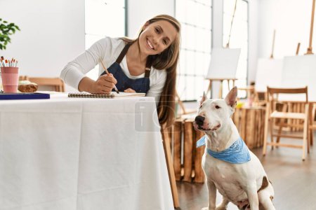 Téléchargez les photos : Jeune femme caucasienne souriant dessin confiant avec chien au studio d'art - en image libre de droit