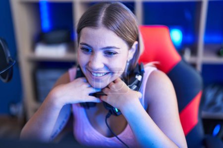 Foto de Joven hermosa mujer hispana streamer sonriendo confiado sentado en la mesa en la sala de juegos - Imagen libre de derechos