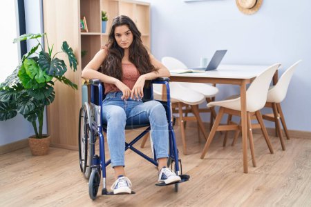 Foto de Young teenager girl sitting on wheelchair at the living room thinking attitude and sober expression looking self confident - Imagen libre de derechos