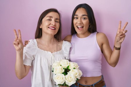 Téléchargez les photos : Hispanic mother and daughter holding bouquet of white flowers smiling with happy face winking at the camera doing victory sign with fingers. number two. - en image libre de droit