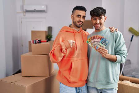 Téléchargez les photos : Jeune couple gay hispanique se déplaçant vers une nouvelle maison tenant des billets pointant du doigt un sourire heureux et fier - en image libre de droit