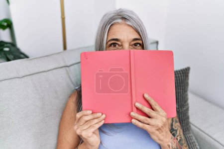 Téléchargez les photos : Femme aux cheveux gris du moyen âge couvrant le visage avec un livre à la maison - en image libre de droit