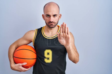 Photo for Young bald man with beard wearing basketball uniform holding ball doing stop sing with palm of the hand. warning expression with negative and serious gesture on the face. - Royalty Free Image