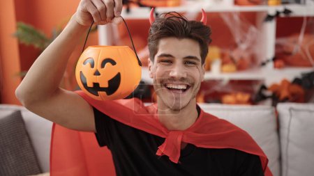 Photo for Young hispanic man wearing devil costume holding halloween pumpkin basket at home - Royalty Free Image