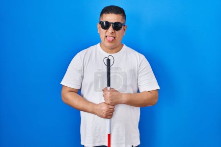 Téléchargez les photos : Hispanic young blind man holding cane sticking tongue out happy with funny expression. - en image libre de droit