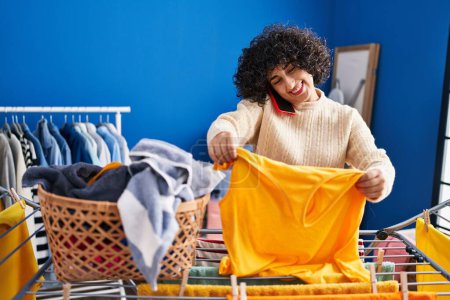 Téléchargez les photos : Young middle east woman talking on the smartphone hanging clothes on clothesline at laundry - en image libre de droit