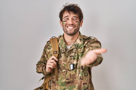 Foto de Joven hispano vistiendo uniforme camuflaje del ejército sonriente alegre ofreciendo mano de palma dando asistencia y aceptación. - Imagen libre de derechos