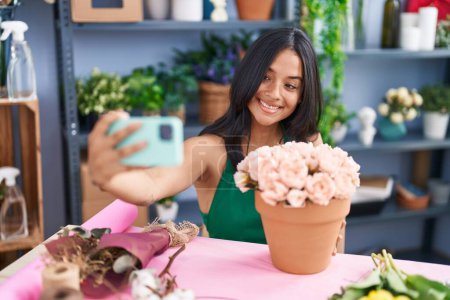 Foto de Young hispanic woman florist make selfie by smartphone sitting on table at florist shop - Imagen libre de derechos