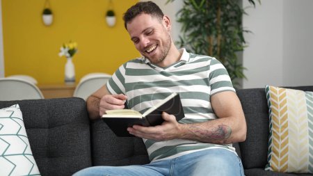 Téléchargez les photos : Young caucasian man reading a book sitting on the sofa at home - en image libre de droit