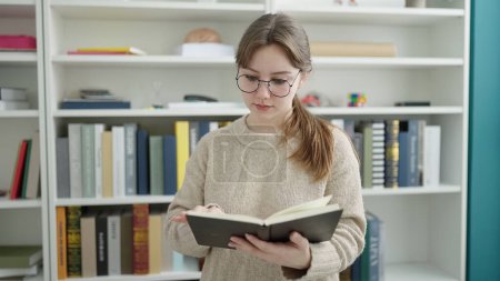 Téléchargez les photos : Jeune femme blonde étudiant livre de lecture à la bibliothèque universitaire - en image libre de droit
