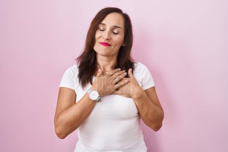 Téléchargez les photos : Middle age brunette woman standing over pink background smiling with hands on chest with closed eyes and grateful gesture on face. health concept. - en image libre de droit