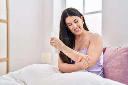Téléchargez les photos : Young hispanic woman applying skin treatment sitting on bed at bedroom - en image libre de droit