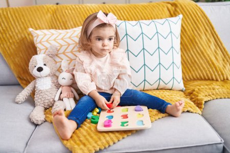Téléchargez les photos : Adorable bambin blonde jouer avec des mathématiques jeu de puzzle assis sur le canapé à la maison - en image libre de droit