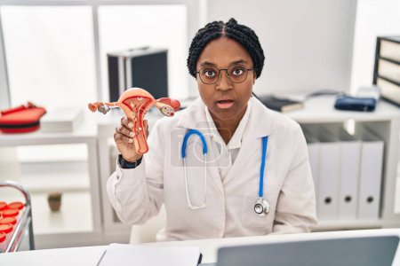 Téléchargez les photos : Médecin afro-américain femme tenant le modèle anatomique de l'organe génital féminin effrayé et étonné avec la bouche ouverte pour la surprise, visage incrédule - en image libre de droit