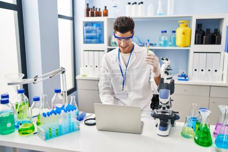 Photo for Young hispanic man wearing scientist uniform using laptop holding test tube at laboratory - Royalty Free Image