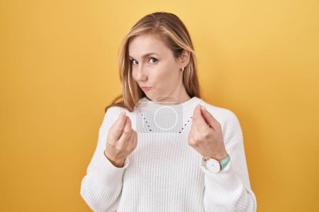 Foto de Young caucasian woman wearing white sweater over yellow background doing money gesture with hands, asking for salary payment, millionaire business - Imagen libre de derechos