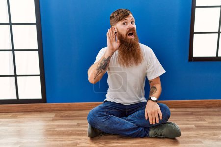 Téléchargez les photos : Homme rousse à longue barbe assis sur le sol dans une pièce vide souriant avec la main sur l'oreille en écoutant une rumeur ou des commérages. concept de surdité. - en image libre de droit