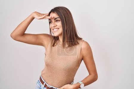 Foto de Young hispanic woman standing over white background very happy and smiling looking far away with hand over head. searching concept. - Imagen libre de derechos