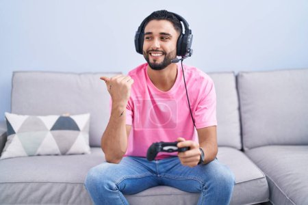 Photo for Hispanic young man playing video game holding controller sitting on the sofa smiling with happy face looking and pointing to the side with thumb up. - Royalty Free Image